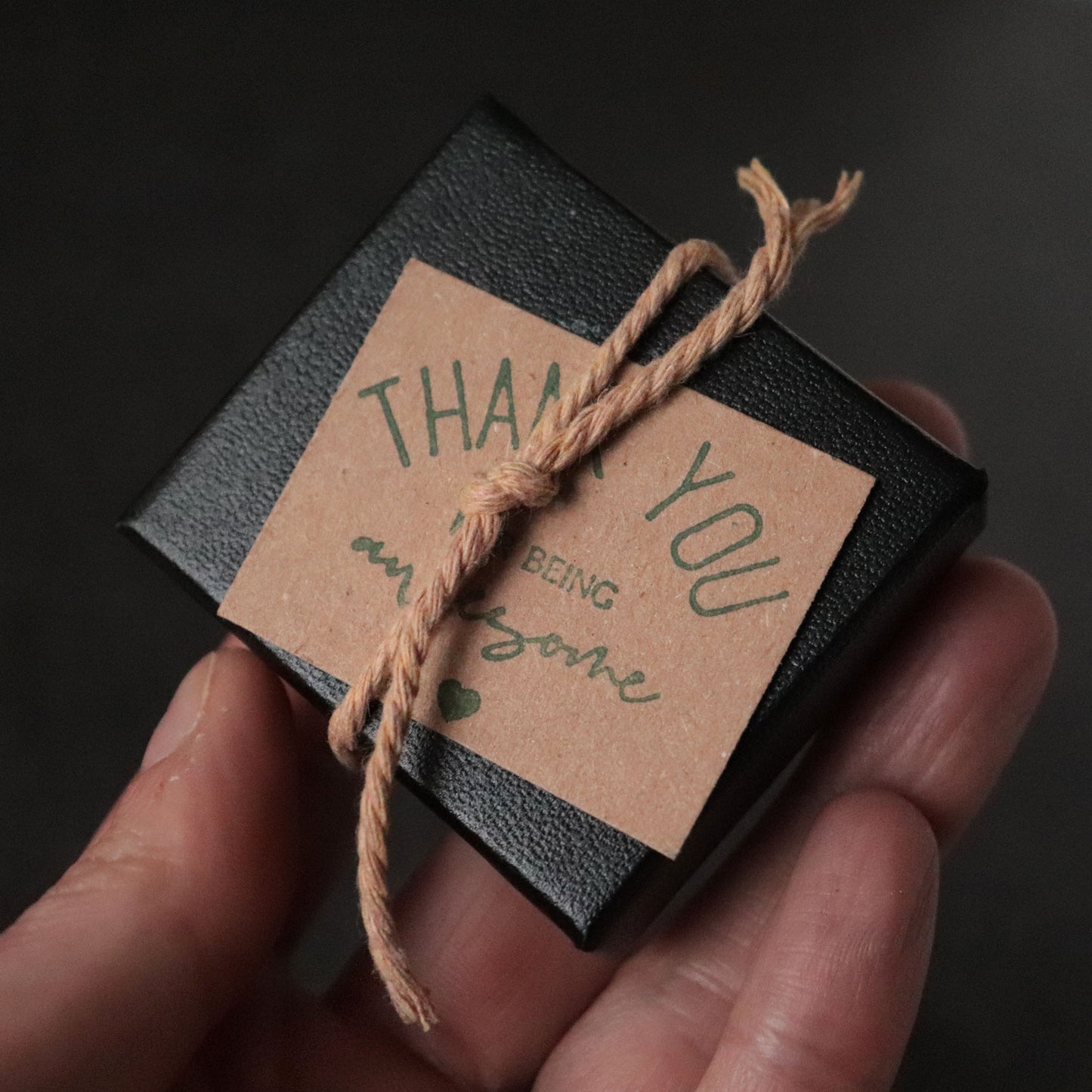 Small black box with a brown tag reading 'Thank you for being awesome' held by a hand against a dark background.