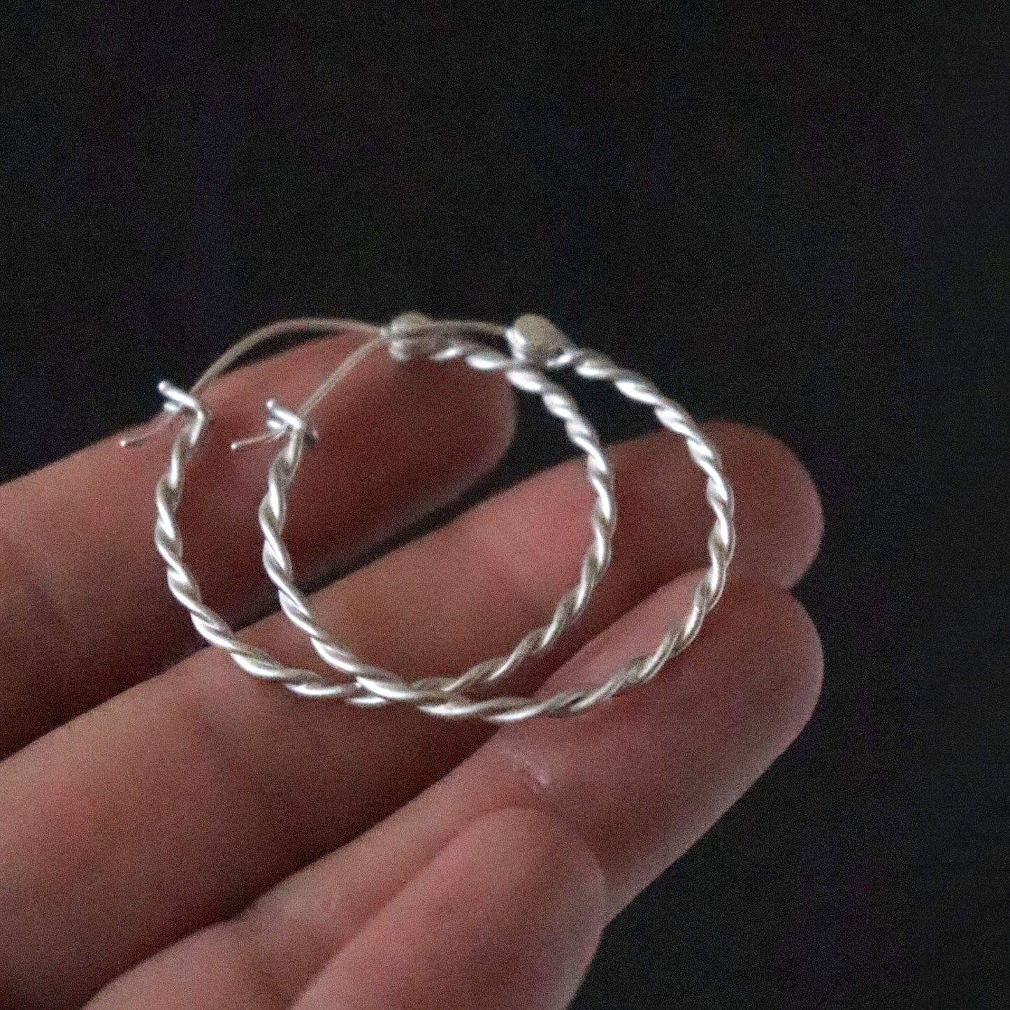 Twisted Silver hoop earrings held in a hand against a dark background