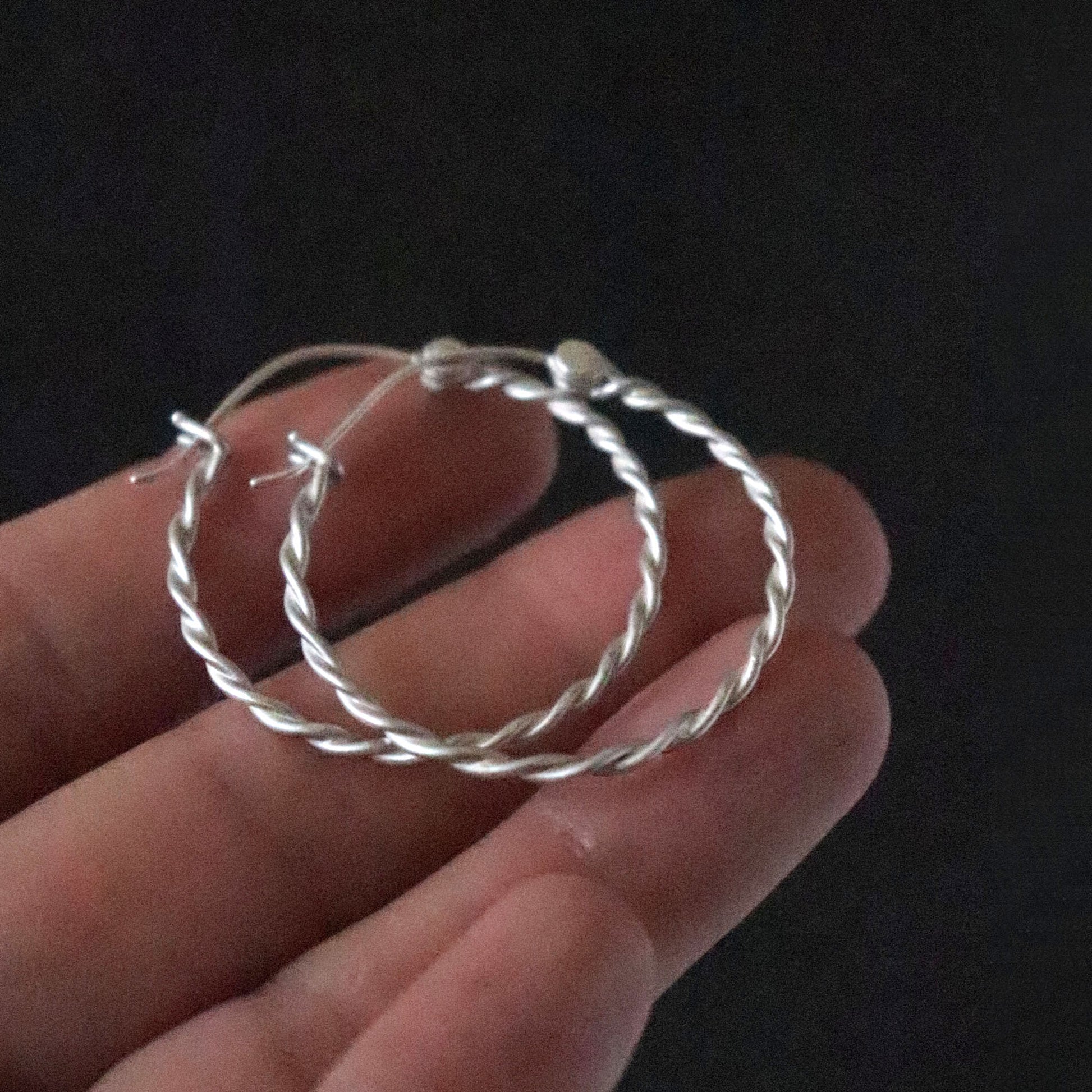 Twisted Silver hoop earrings held in a hand against a dark background