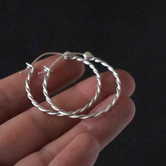 Twisted Silver hoop earrings held in a hand against a dark background