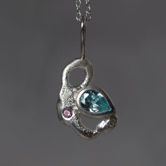 Silver pendant with a blue pear shape gemstone and pink stone on a dark gray background