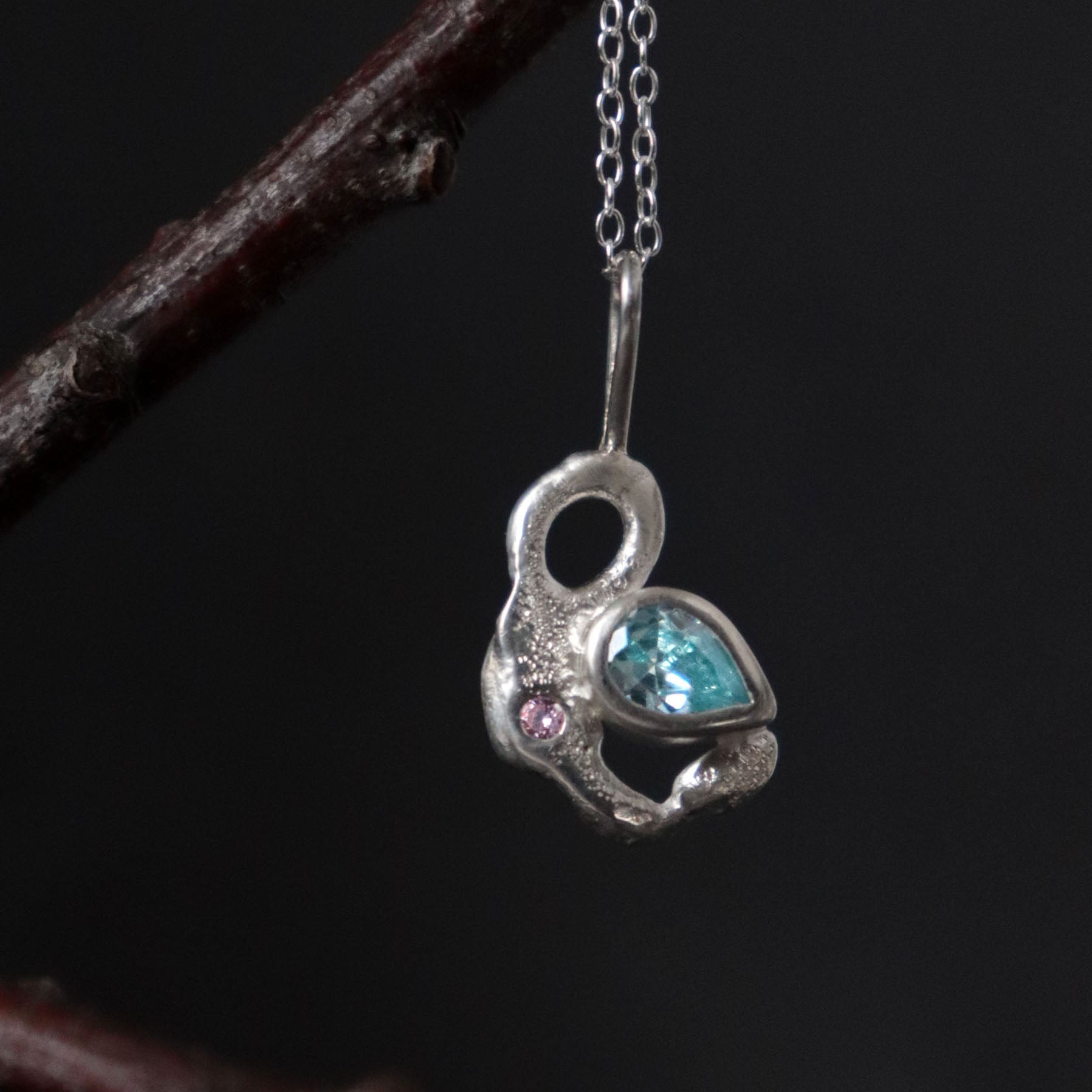 Silver necklace with a organic shape pendant featuring blue and pink stones on a dark background.